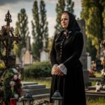 Žena v tradiční černé smuteční róbě u starého železného kříže na hřbitově za šera